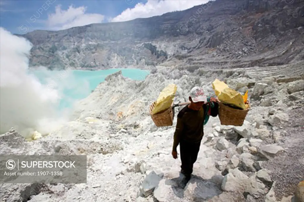 Sightseeing on the lake of the crater of the Kawa Ijan vulcano where a sulphur mine constantly smokes at the foreground a sulphur worker carrying 80 kg of sulphur Baluran national park Java Indonesia