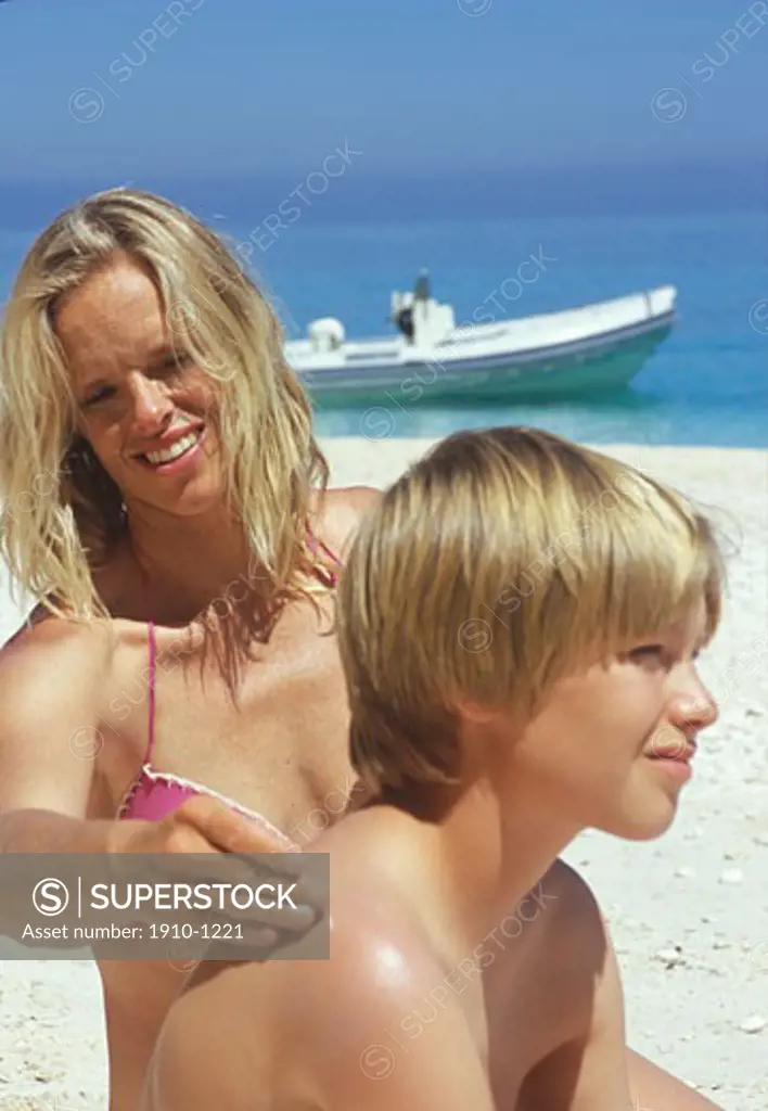 Mother lathers sunscreen on sons 12 yrs back  beach and inflatable boat in sea behind  Italy  Sardinia
