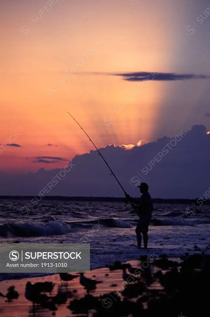 A surf fisherman is silhouetted by the setting sun