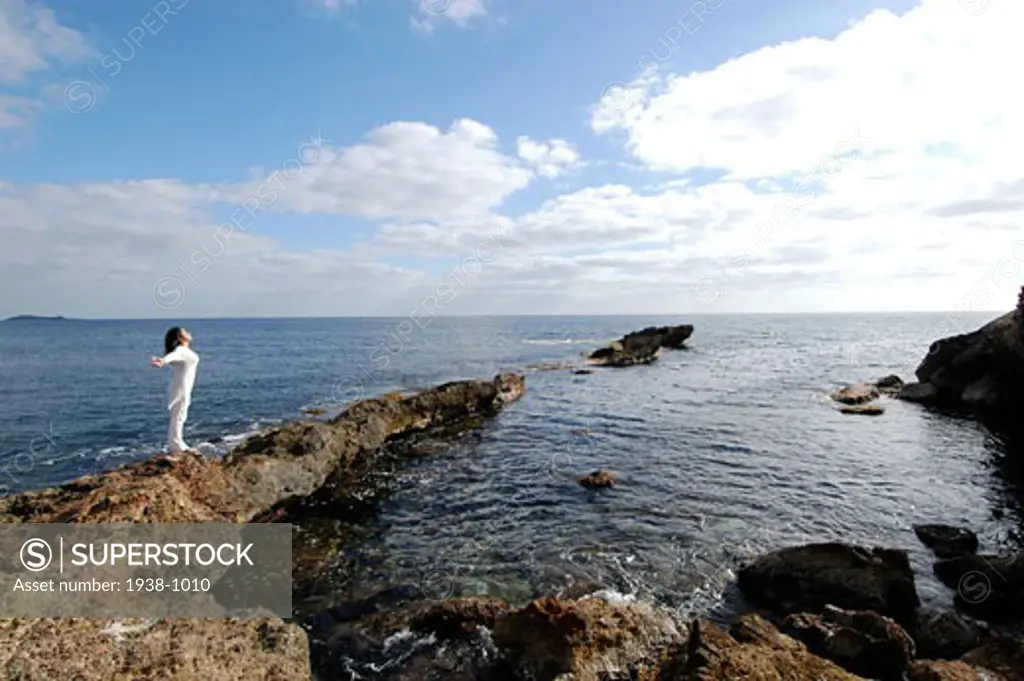 Mixed race young woman  half asian half spanish  standing on the rocks next to the ocean  breathing and relaxing  in Ibiza  Spain