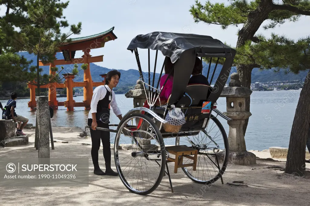 Smiling runner talking with tourists seated in his rickshaw on Miyajima Island, Japan. In the background is the gate, or torii, marking the entrance t...