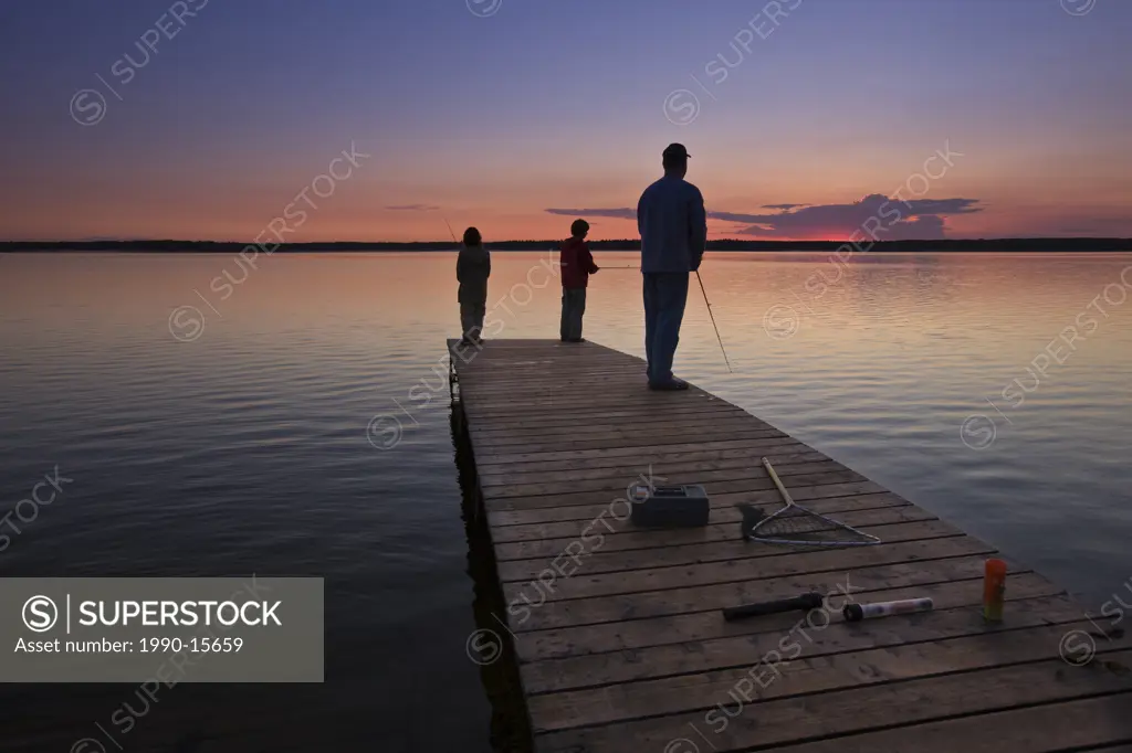 A father with his two sons fishing at the end of a wharf on Lake Audy at sunset, Riding Mountain National Park, Manitoba, Canada