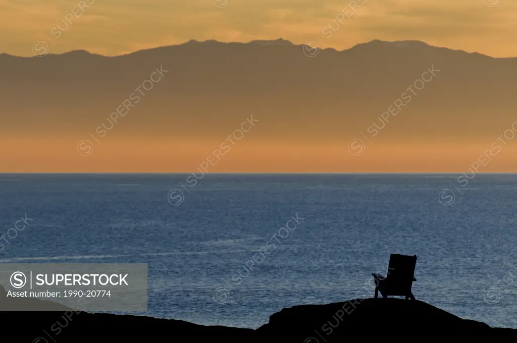 An adirondack_style chair is silhouetted against the ocean and the Olympic Mountains at sunset near Victoria BC.