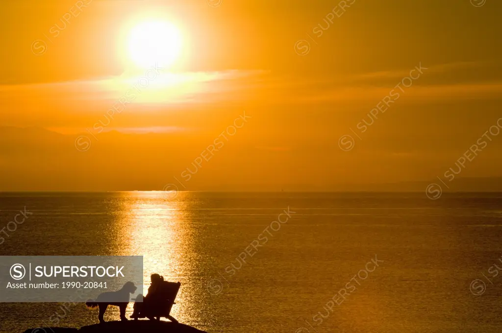 A woman and her dog are silhouetted by the sunset on an oceanside rock off Beach Drive, Victoria BC.
