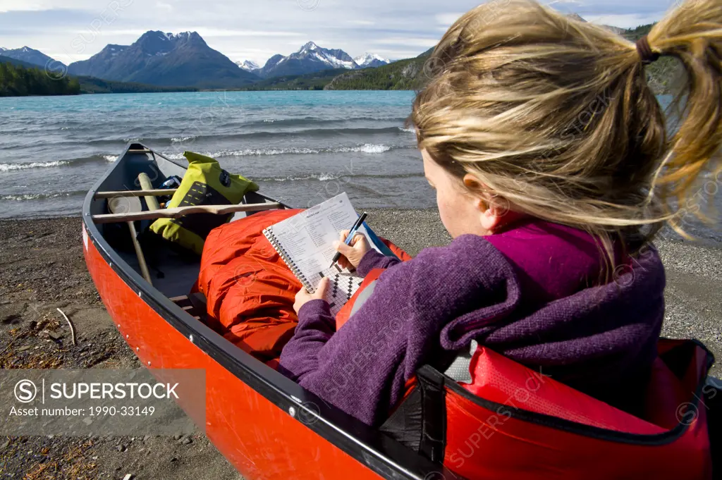 A young, attractive woman does a crossword puzzle sitting in a canoe on the shores of the azure Tatlayoko Lake, British Columbia, Canada