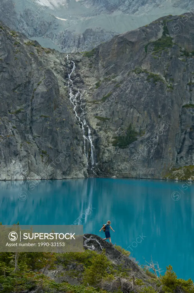 A young woman hikes in front of a blue lake and waterfall in the Niut Range, British Columbia, Canada