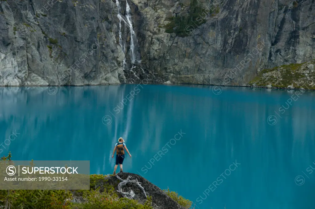 A young woman hikes in front of a blue lake and waterfall in the Niut Range, British Columbia, Canada