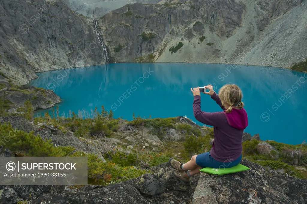 A young woman snaps a photo of a waterfall and blue lake with a point_and_shoot camera, British Columbia, Canada