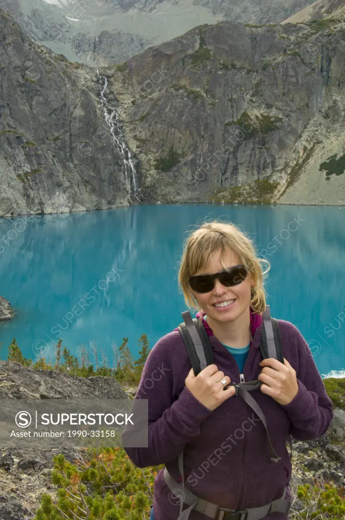 A young, attractive woman with sunglasses smiles in front of a turquoise blue lake and waterfall in the Coast Mountains, British Columbia, Canada