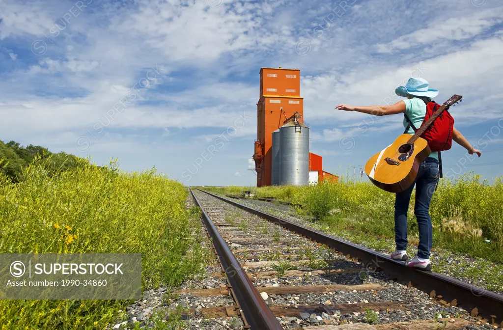 18 year old girl with guitar walking along railway with abandoned grain elevator in the background, Carey, Manitoba, Canada