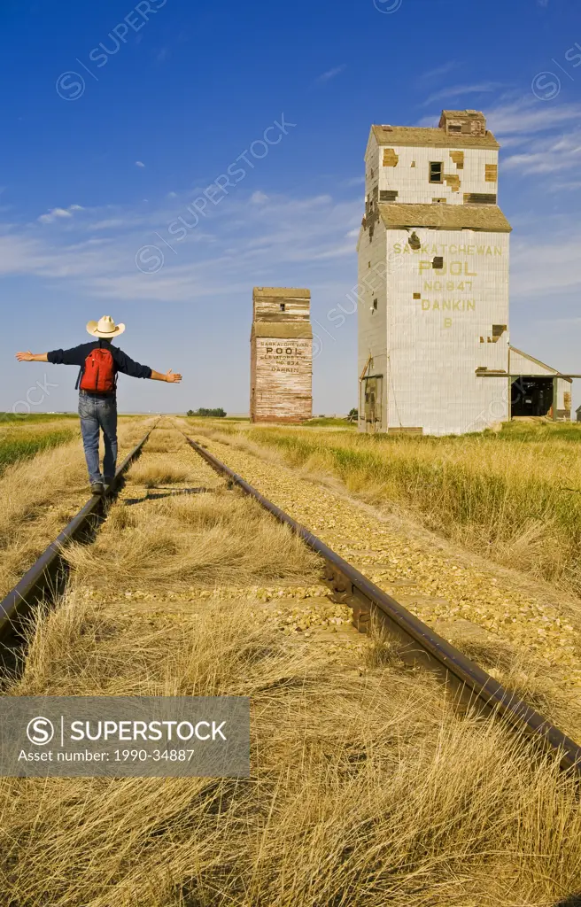 a man hikes along the railway with abandoned elevators in the background, Dankin, Saskatchewan, Canada