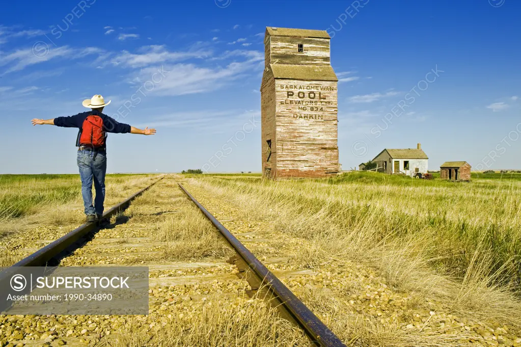 a man hikes along the railway with abandoned elevators in the background, Dankin, Saskatchewan, Canada