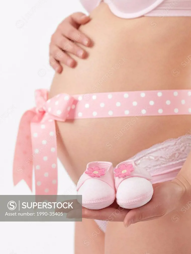 Pregnant woman with a pink bow on her belly holding baby shoes in her hand