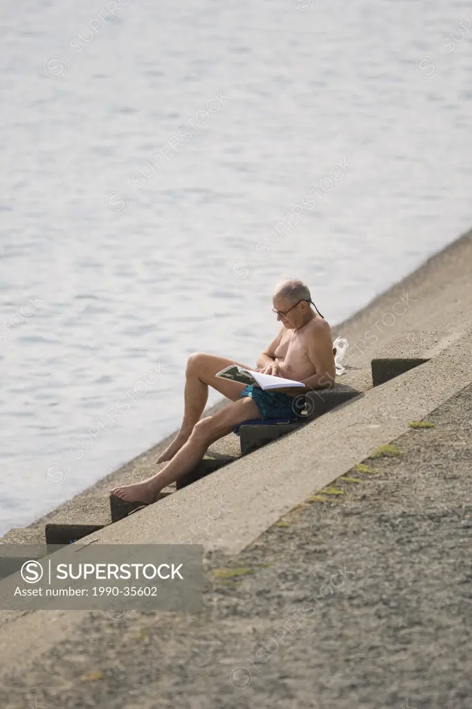 A senior citizen soaks up the sun at Westwood Lake while reading a book. Nanaimo, Vancouver Island, British Columbia, Canada.