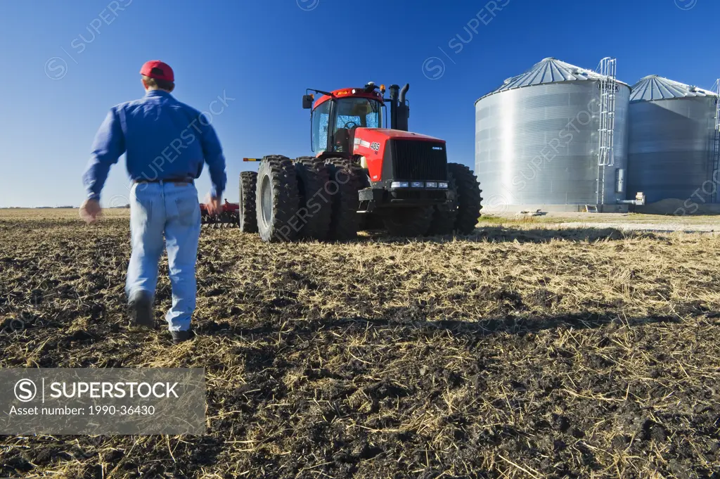 a man walks through newly cultivated soil and wheat stubble towards a tractor pulling cultivating equipment and grain storage bins in the background, ...
