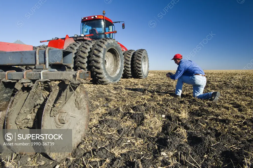 a man examines newly cultivated soil and wheat stubble beside a tractor pulling cultivating equipment near Lorette, Manitoba, Canada