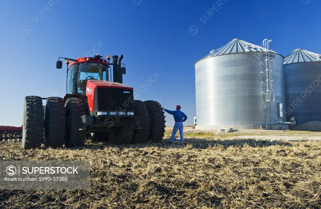 a man next to a tractor pulling cultivating equipment loks out over a newly cultivted field with grain storage bins in the background, near Lorette, M...