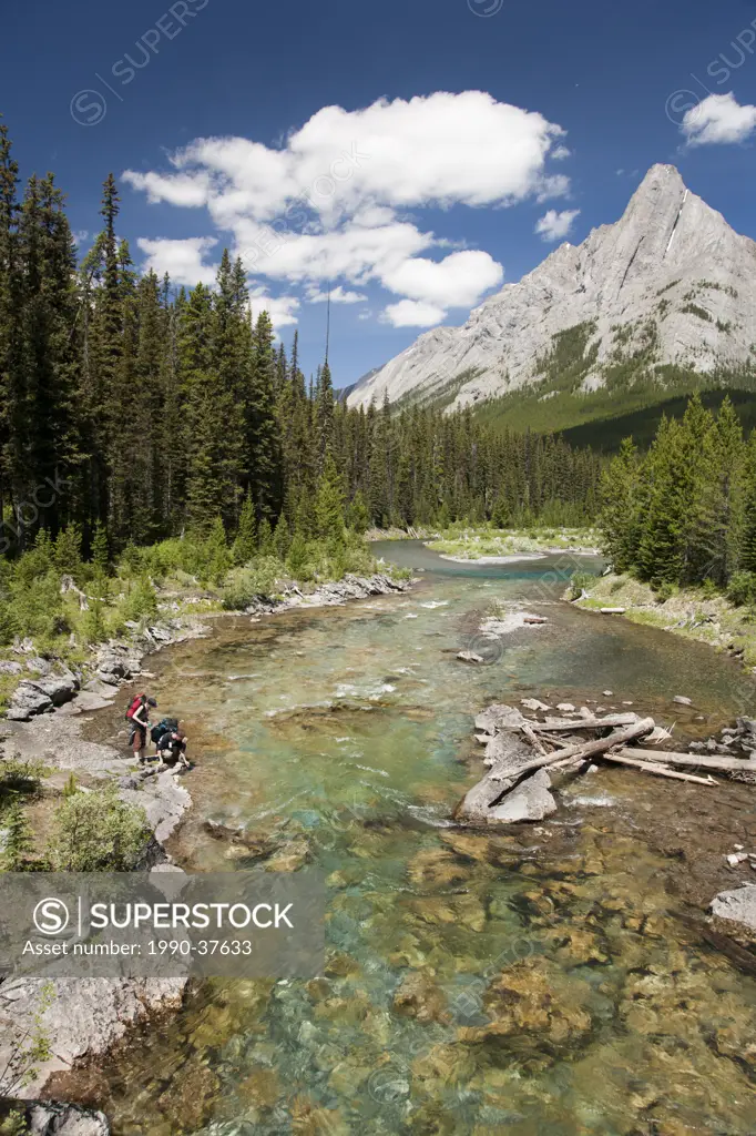 Two hikers collecting water on Bryant Creek with Cone Mountain in the background, Banff National Park, Alberta, Canada.