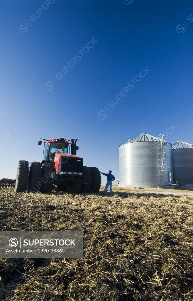 a man next to a tractor pulling cultivating equipment looks out over a newly cultivted field with grain storage bins in the background, near Lorette, ...