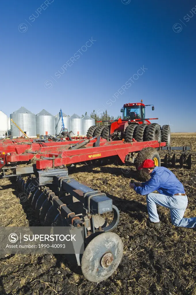 a man holds newly cultivated soil and wheat stubble next to a tractor pulling cultivating equipment, grain storage bins in the background, near Lorett...