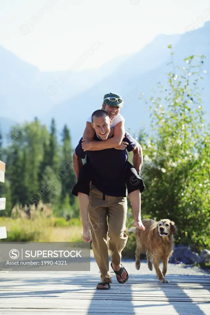A fun-loving couple runs across a pedestrian bridge in the mountains with the husband piggybacking the wife and the family dog in tow, Canada