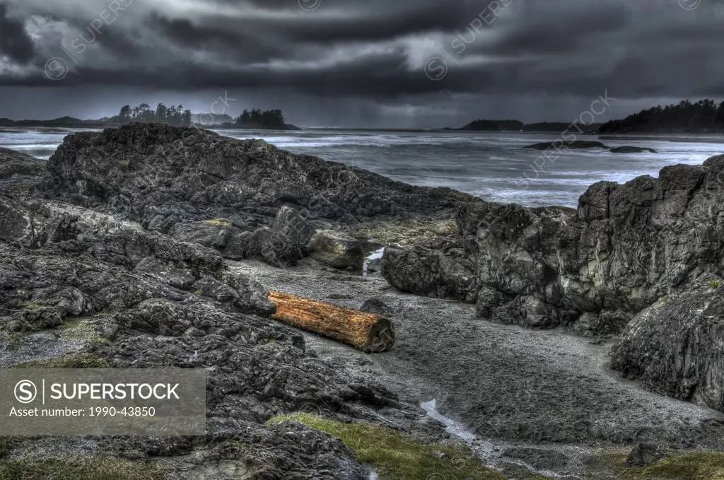 Dark stormy Tofino seascape with rocks and log in foreground, islands in back.