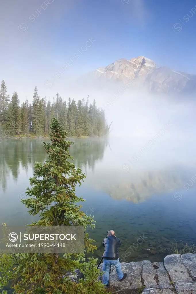 Photographer on shore of Pyramid Lake with Pyramid Mountain partly obscured by mist, Jasper National Park, Alberta, Canada