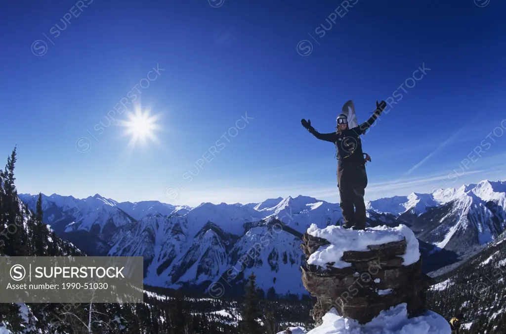 Snowboarder standing on boulder overlooking mountain range in Backcountry of Alberta, Canada.