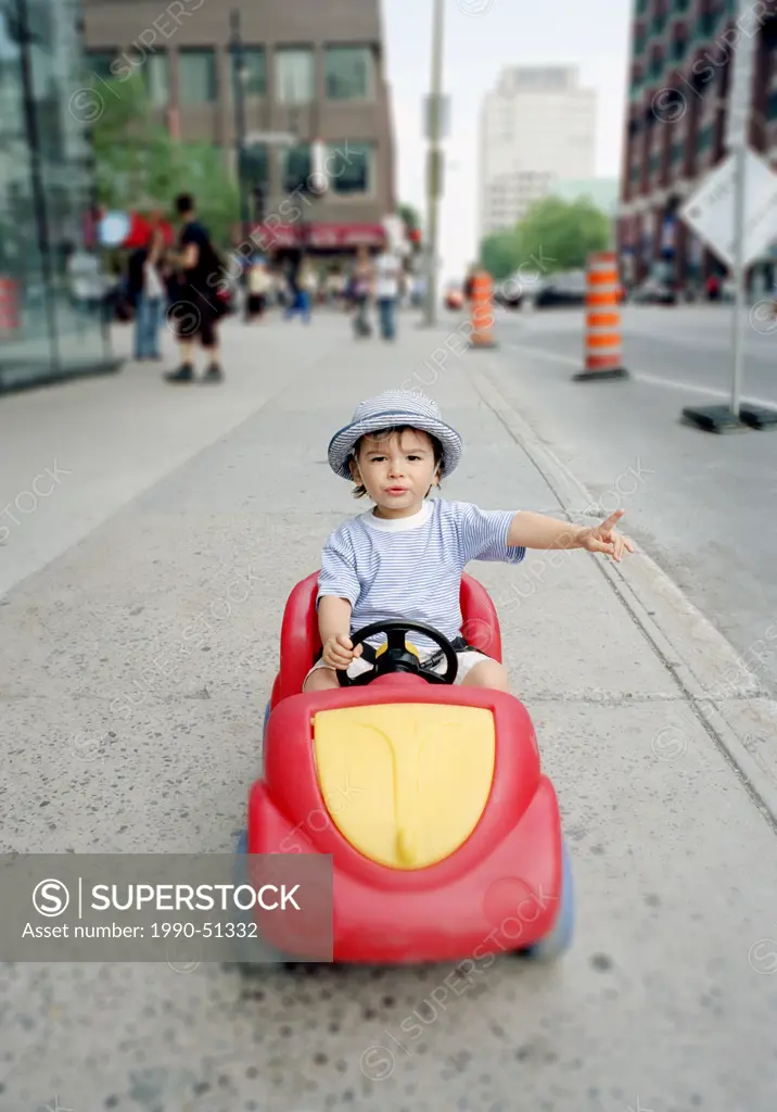 twenty month old eurasian boy / child in a toy plastic car on the sidewalk dowtown city center with selective focus, Montreal, Quebec, Canada.