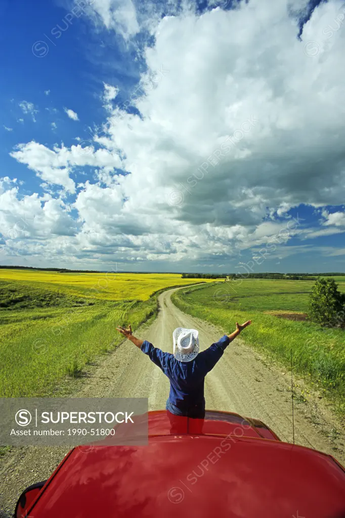 Man sits on the front of apick_up a truck on a country road with farmland in the background, Tiger Hills, Manitoba, Canada