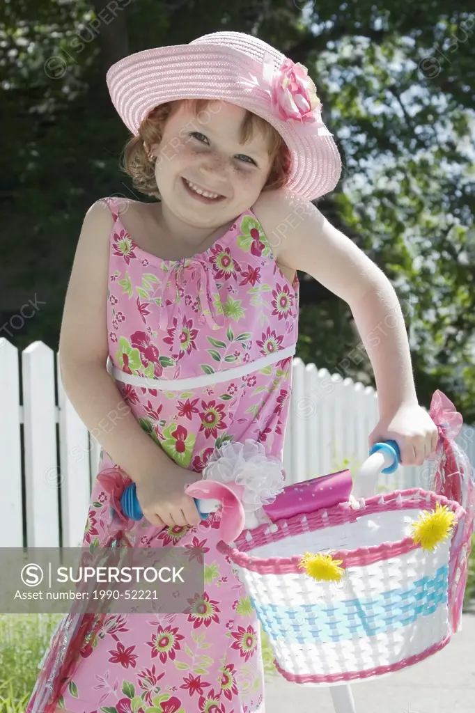 Girl with sundress and hat standing in front of picket fence with bicycle, Canada.