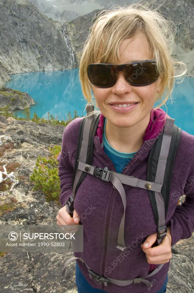 A young, attractive woman with sunglasses smiles in front of a turquoise blue lake and waterfall in the Coast Mountains, British Columbia, Canada