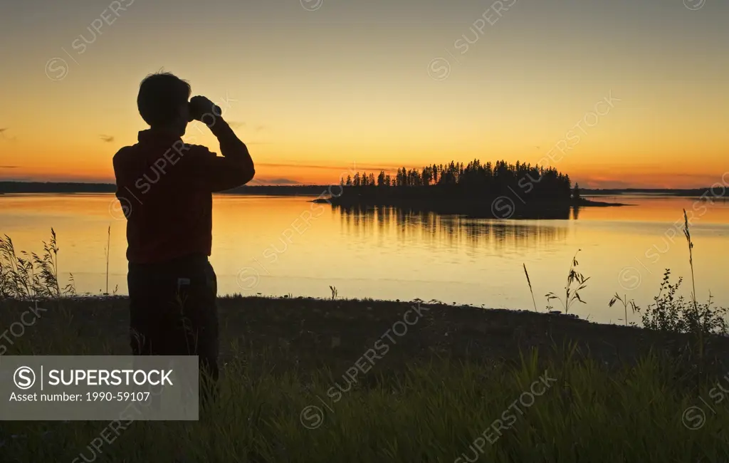 Middle age male hiker looking out over Astotin Lake with binoculars at dusk, Elk Island Park, Alberta, Canada.