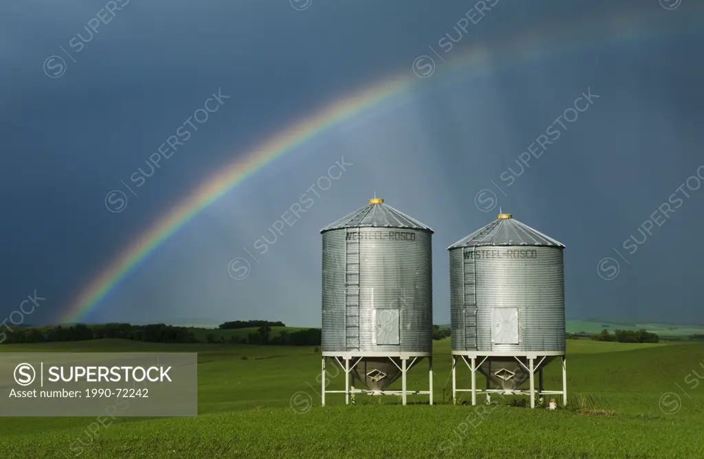 Grain field with old grain hopper bins(silos) and rainbow in the background, Tiger Hills, Manitoba, Canada