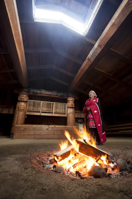 A first nations elder dressed in her traditional regalia stands beside a fire in the bighouse in the village of Oweekeno. Wuikinuxv, Rivers Inlet, Bri...
