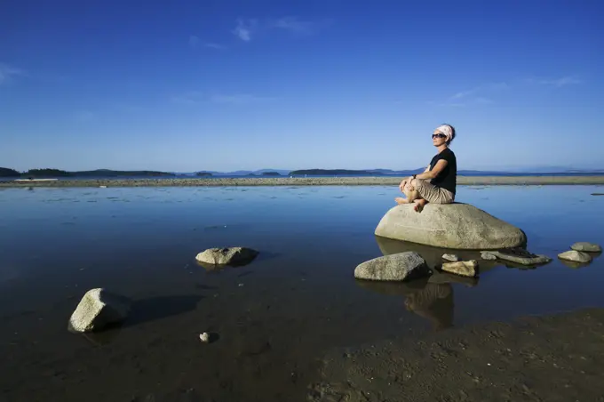 A mature woman relaxes amongst rocks at low tide while visiting Sidney Spit.  Gulf islands national park reserve,  Vancouver Island, British Columbia, Canada