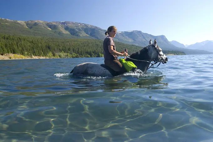 A young woman and her horse wade in the shallow, clear waters of Tatlayoko Lake, British Columbia, Canada