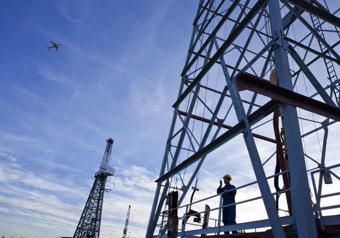 Drilling rig worker talking on radio on platform with airplane flying overhead, Alberta, Canada