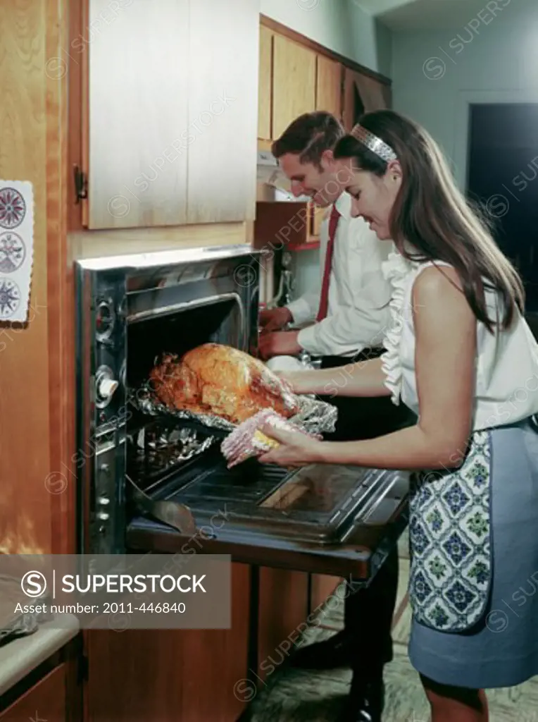 Side profile of a young woman taking a roasted turkey out of an oven with a young man smiling