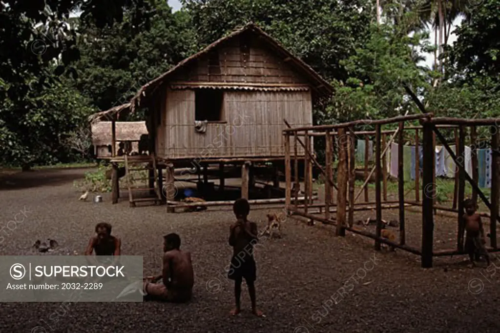 Two men sitting outside a stilt house with a boy nearby, Milne Bay, Papua New Guinea