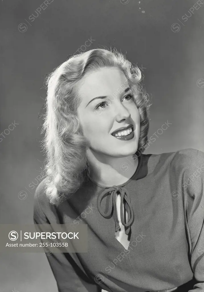Blonde woman with wavy hair wearing blouse with tie in front smiling looking up at corner