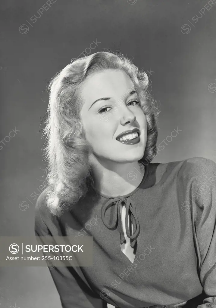 Blonde woman with wavy hair wearing blouse with tie in front smiling with chin raised