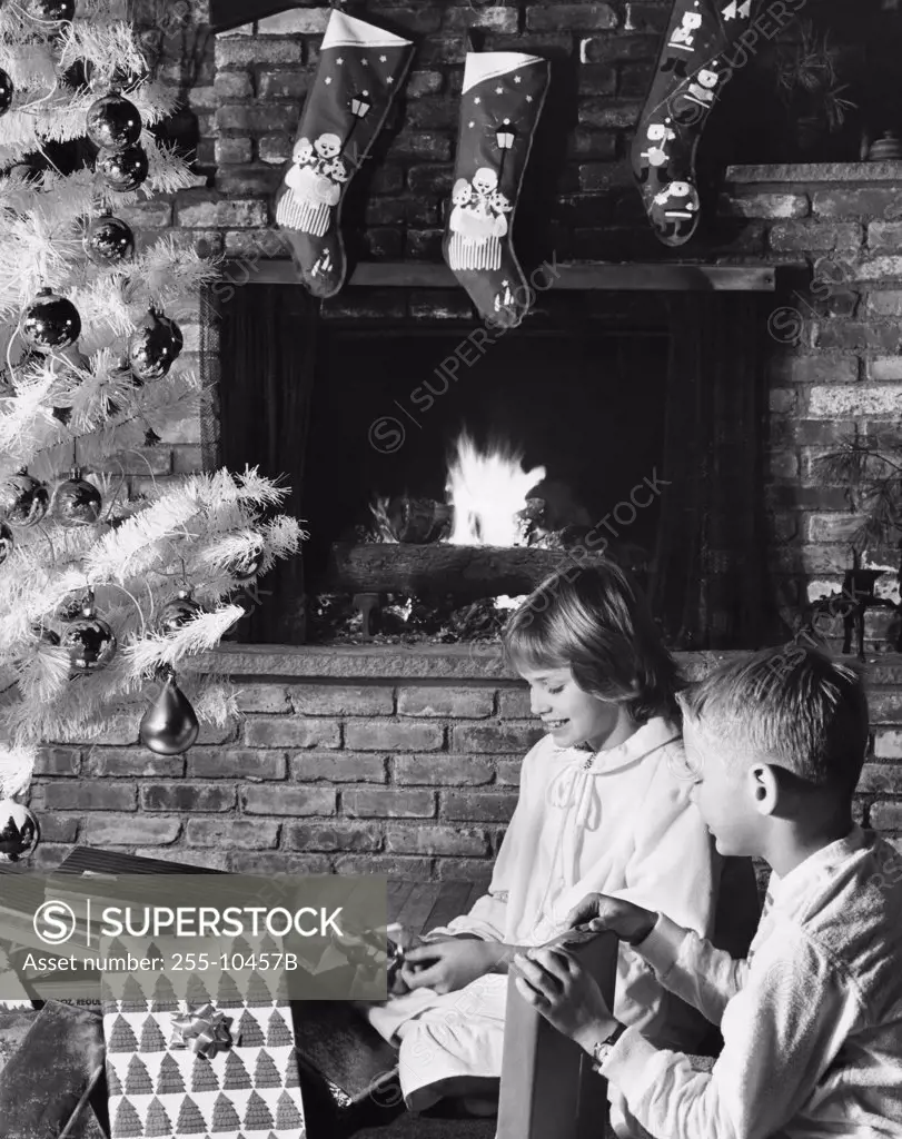 Side profile of a girl and her brother unwrapping Christmas presents near a fireplace