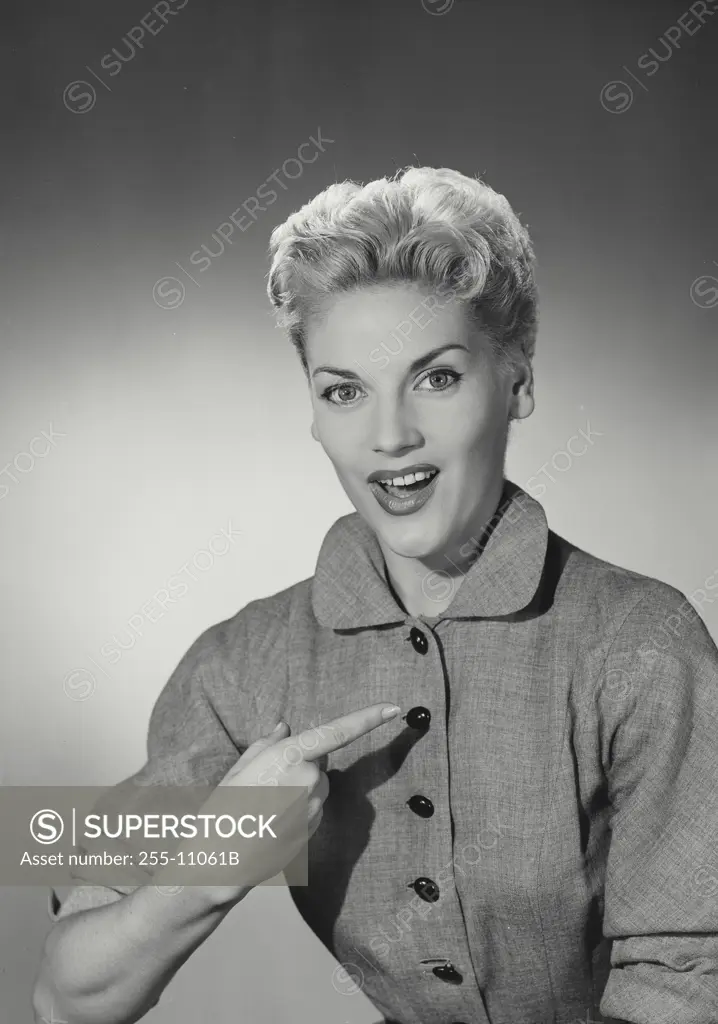 Vintage Photograph. Smiling blonde woman with hair up wearing coat with black buttons point finger back at chest