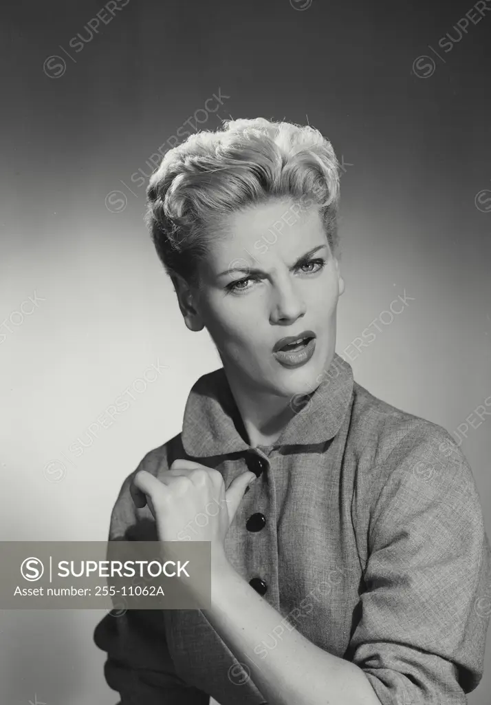 Vintage Photograph. Blonde woman with hair up wearing coat with black buttons gesturing thumb back at chest