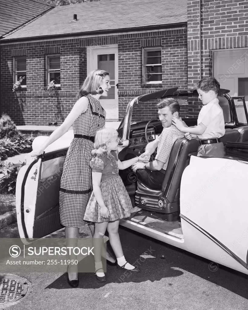 Mid adult woman and her daughter standing beside a car with a mid adult man and his son sitting in a car