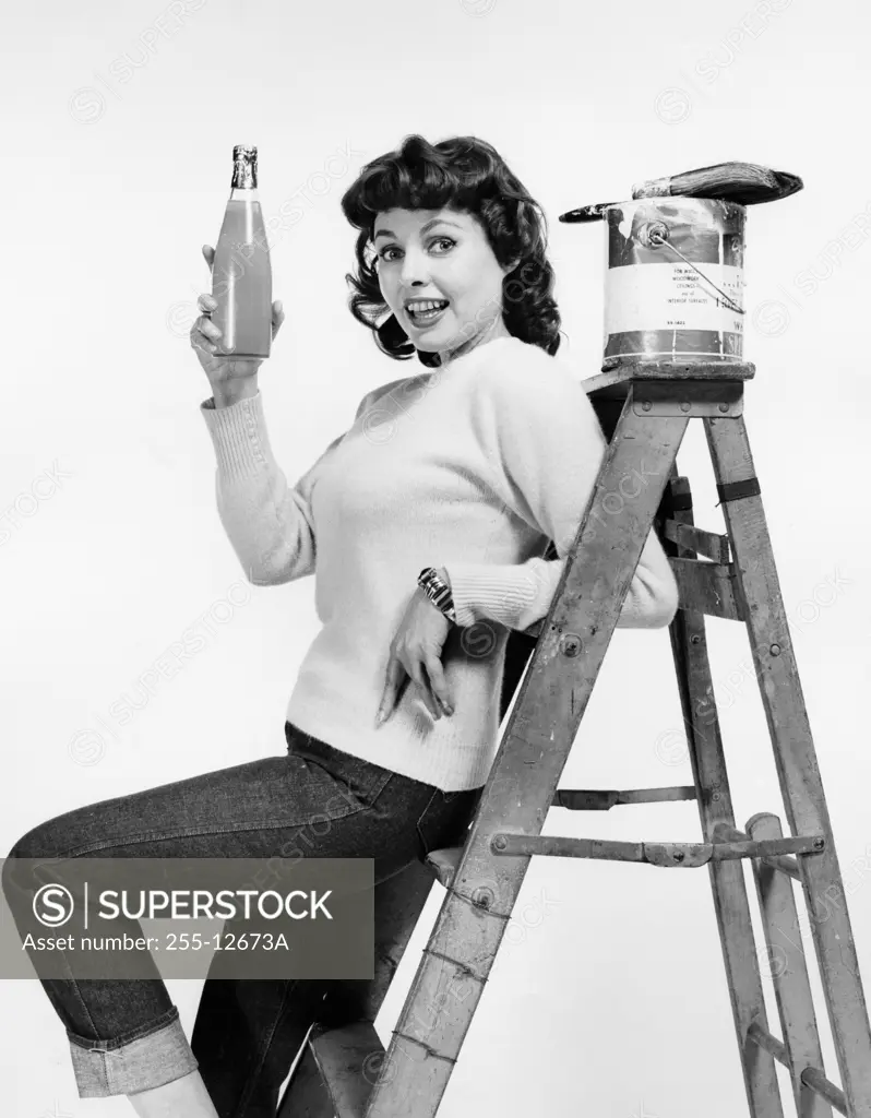 Portrait of a young woman leaning against a step ladder and holding a bottle