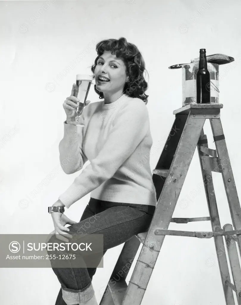 Portrait of a young woman sitting on a step ladder and drinking beer