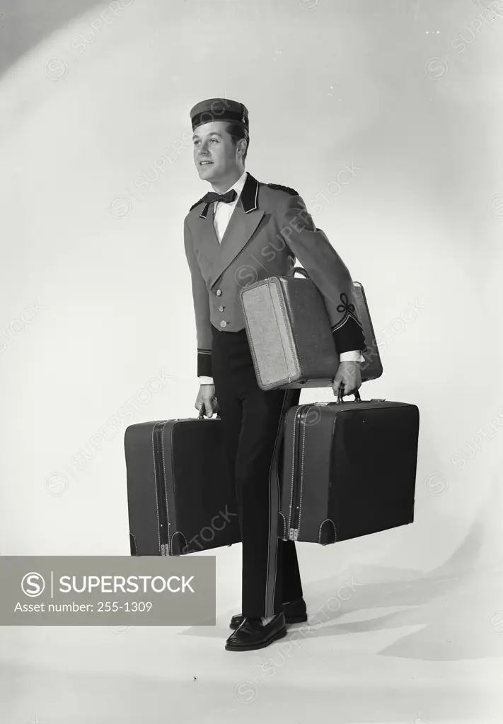 Vintage Photograph. Man wearing bellman uniform turned to side on white background holding three suitcases