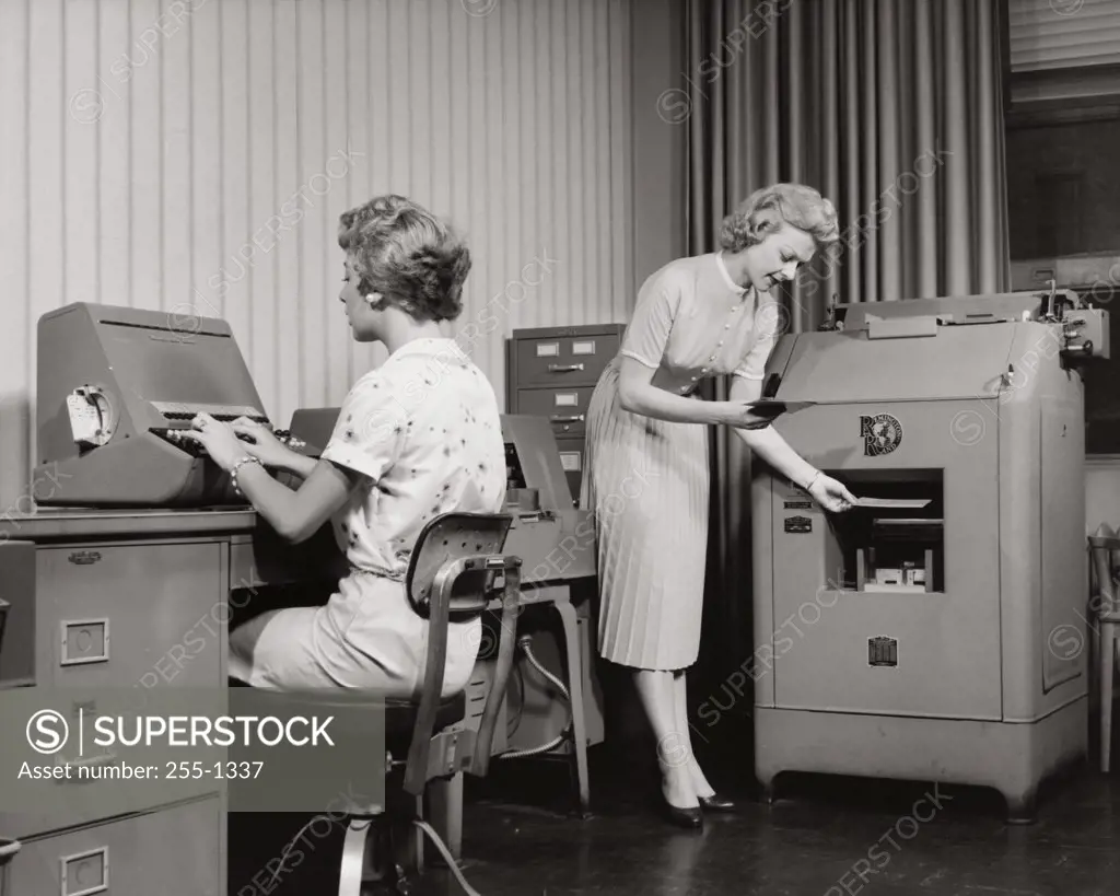 Two mid adult women working on a punch card tabulating system in an office